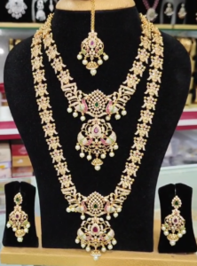 22K gold-plated royal bridal temple necklace set with ruby, emerald, zirconia accents, pearls, and two necklaces (short and long), featuring intricate designs and matching earrings.
